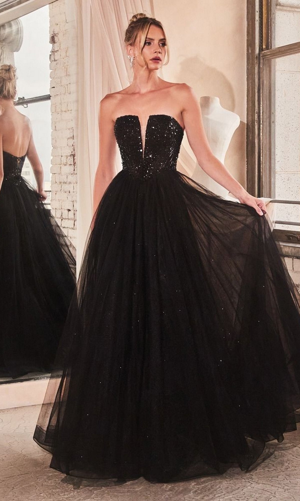 A woman stands indoors near a window and mirror, wearing the Lace-Up Strapless Long Glitter Prom Dress CD0217 featuring a floor-length black gown with a deep neckline and full tulle skirt.